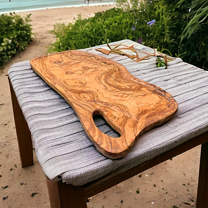 Oblong olive wood cutting board with juice groove, beautiful wood grain, fluid line, and grip handle. Size: 23-1/2 x 11 x 1/2 inches. Versatile as a presentation board or charcuterie board. Rustic elegance with rich vibrant color, organic look and feel, blending harmoniously with the outdoors.