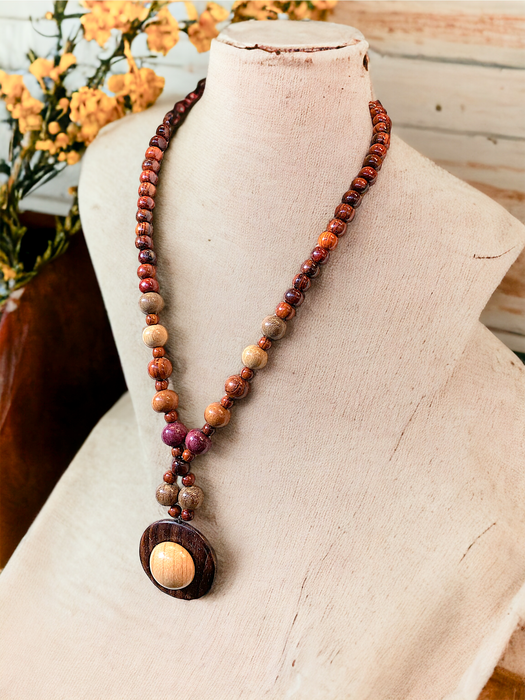 Handmade Necklace Elizabeth in assorted exotic woods, featuring a round pendant, displayed on a bust with flowers in the background.
