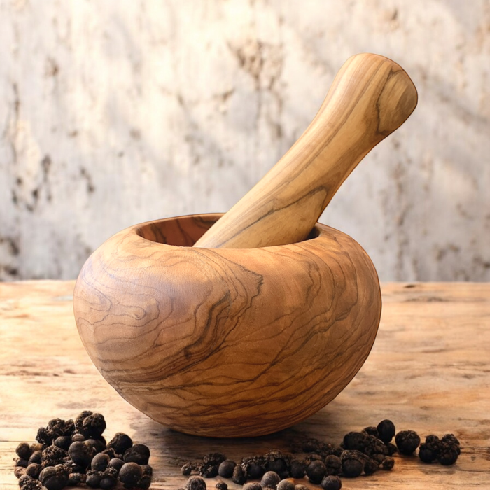 An olive wood mortar and pestle with rounded sides is displayed on a rustic wooden countertop with black peppercorns. 