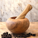 An olive wood mortar and pestle with rounded sides is displayed on a rustic wooden countertop with black peppercorns. 